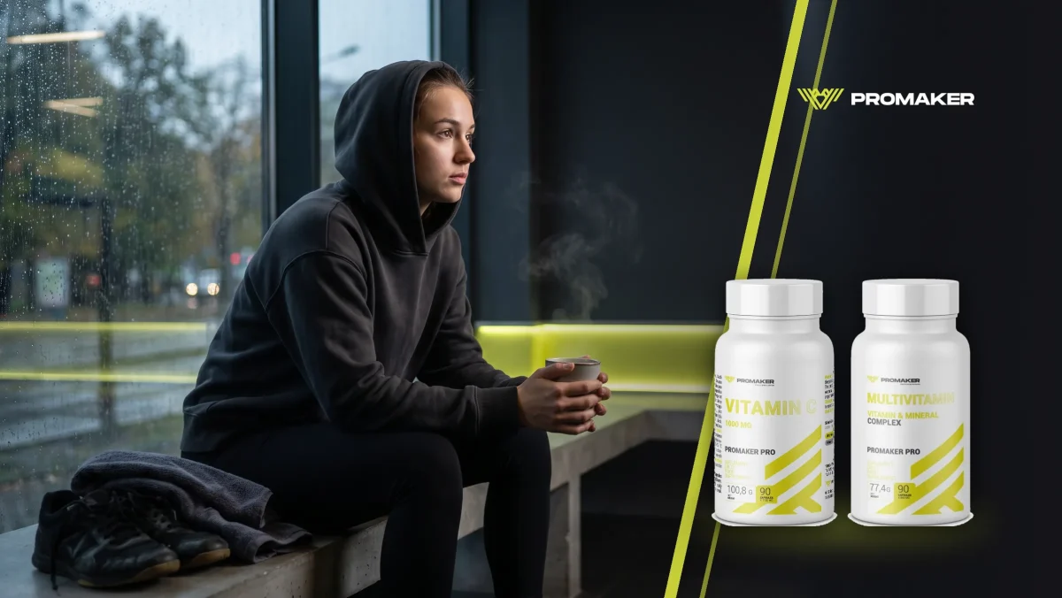 Młoda kobieta w ciemnej bluzie z kapturem siedzi na ławce w siłowni z kubkiem gorącej herbaty, za oknem deszcz; obok Vit C 1000 mg i Multivitamin Complex Promaker