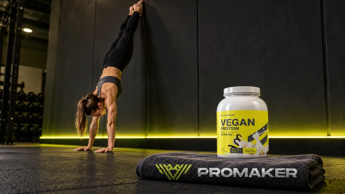 Kobieta w staniu na rękach przy ścianie w siłowni, obok odżywka Vegan Protein PROMAKER i ręcznik z logo