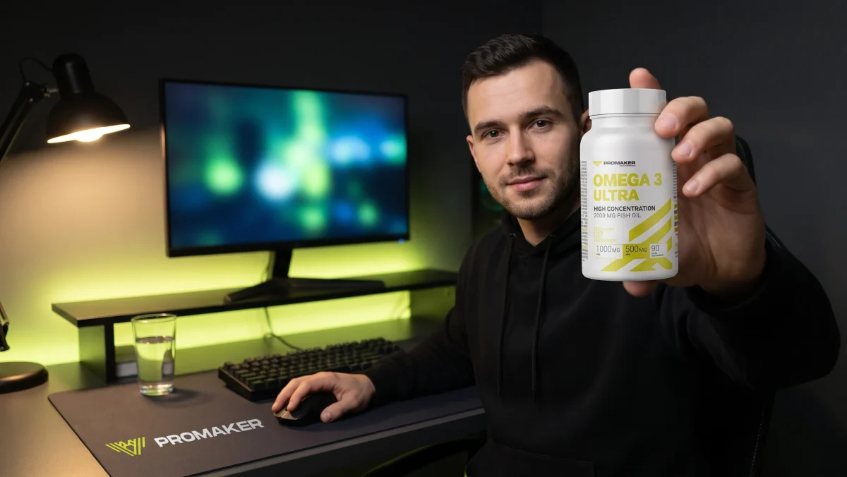 Gracz przy biurku z monitorem trzyma kapsułki Omega ULTRA PROMAKER, na biurku podkładka pod mysz z logo PROMAKER