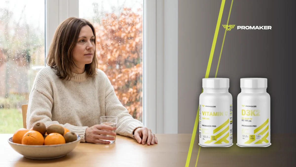Kobieta w kremowym swetrze siedzi przy stole przy oknie w deszczowy jesienny dzień ze szklanką wody, obok miska pomarańczy i kiwi; z prawej Vit C 1000 mg i Vit D3+K2 Promaker
